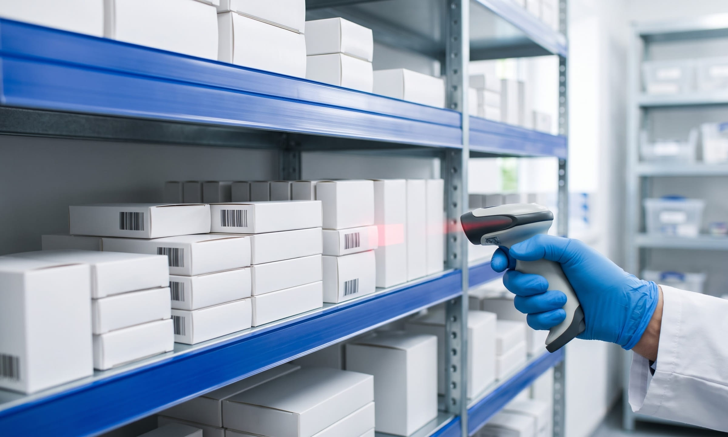 Plain white medicine cartons on clean steel shelving, with a gloved hand scanning one using a handheld barcode scanner.