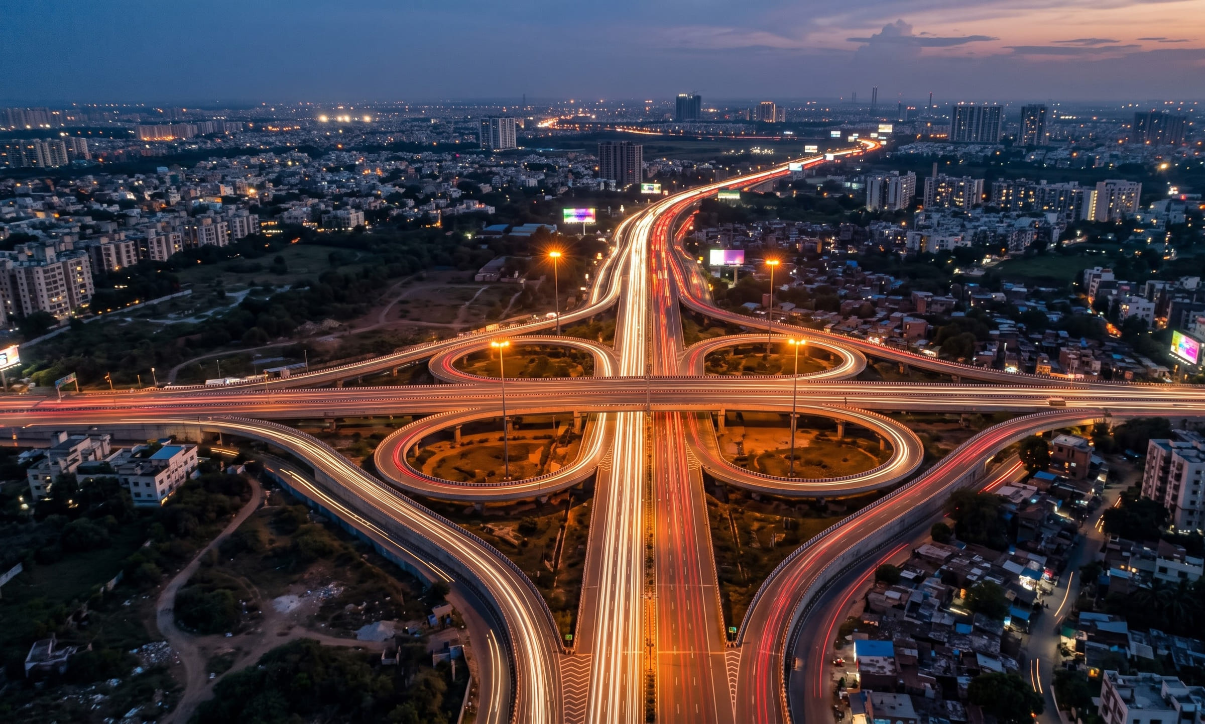 An aerial long exposure of a large highway interchange at dusk, with white and red light trails under a deep-blue sky.